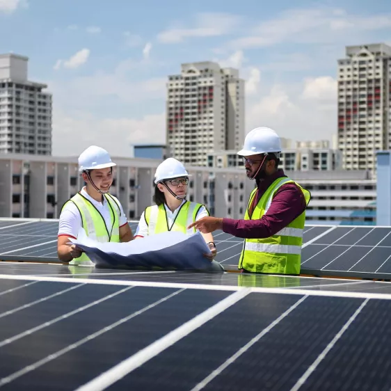 Three people next to solar panels on the rooftop of a building. All wear reflective vests and helmets. One of the people has some sheets in his hands and the three talk about the indications given on those sheets.